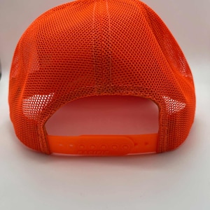 May include: Bright orange trucker hat with a mesh back and adjustable snap closure. The hat features a curved brim and a button on top. The back of the hat has a breathable mesh design.