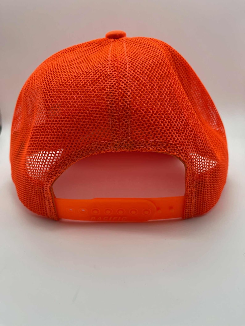 May include: Bright orange trucker hat with a mesh back and adjustable snap closure. The hat features a curved brim and a button on top. The back of the hat has a breathable mesh design.