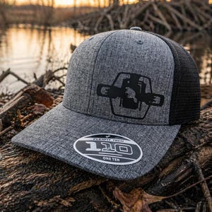 May include: A gray Flexfit Tech 110 hat with a black mesh back and a black graphic on the front. The graphic is a stylized design within a hexagon shape. The hat has a curved brim and a Flexfit Tech 110 sticker.