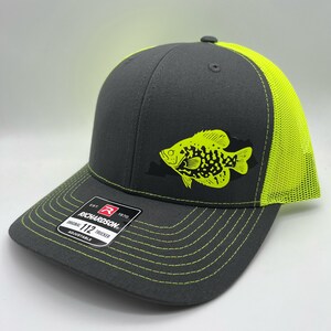 May include: A gray Richardson 112 trucker hat with a neon green fish graphic. The hat features a neon green mesh back and brim stitching. The front panel displays the Richardson logo and "Original 112 Trucker" text.