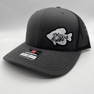 May include: Charcoal gray trucker hat with a black mesh back and a white and black fish patch. The hat has a curved brim and an adjustable strap. The Richardson 112 hat is adjustable.