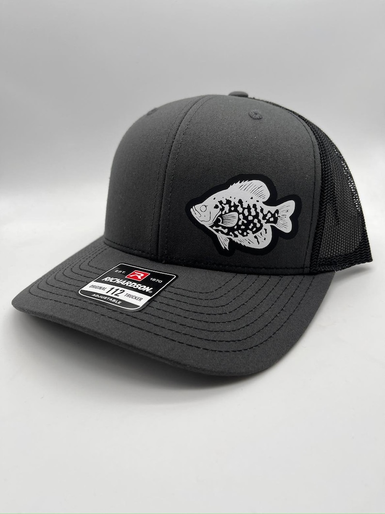 May include: Charcoal gray trucker hat with a black mesh back and a white and black fish patch. The hat has a curved brim and an adjustable strap. The Richardson 112 hat is adjustable.