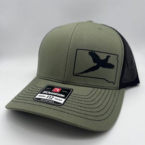 May include: Olive green trucker hat with a black mesh back and a curved brim. The front features a black graphic of a bird inside a state outline. The hat has a Richardson 112 Trucker label.