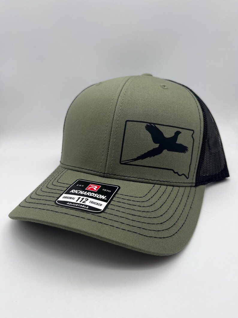 May include: Olive green trucker hat with a black mesh back and a curved brim. The front features a black graphic of a bird inside a state outline. The hat has a Richardson 112 Trucker label.