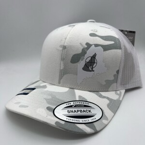 Pode incluir: Bon&eacute; de caminhoneiro branco camuflado com uma silhueta de lobo branco dentro do contorno do estado do Maine. O bon&eacute; tem um s&iacute;mbolo de alvo preto e branco no lobo. O bon&eacute; tem um adesivo preto e branco no lado que diz "The Authentic Yupoong Snapback Yupoong Since 1974."