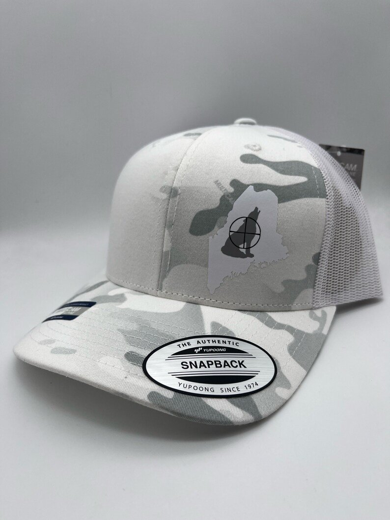 Pode incluir: Bon&eacute; de caminhoneiro branco camuflado com uma silhueta de lobo branco dentro do contorno do estado do Maine. O bon&eacute; tem um s&iacute;mbolo de alvo preto e branco no lobo. O bon&eacute; tem um adesivo preto e branco no lado que diz "The Authentic Yupoong Snapback Yupoong Since 1974."