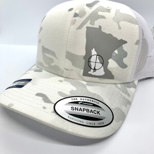 Pode incluir: Bon&eacute; de caminhoneiro branco camuflado com um contorno cinza de Minnesota e uma silhueta de lobo dentro de um alvo. O bon&eacute; tem uma malha branca na parte de tr&aacute;s e um adesivo ovalado prateado que diz "The Authentic Yupoong Snapback Yupoong Since 1974".