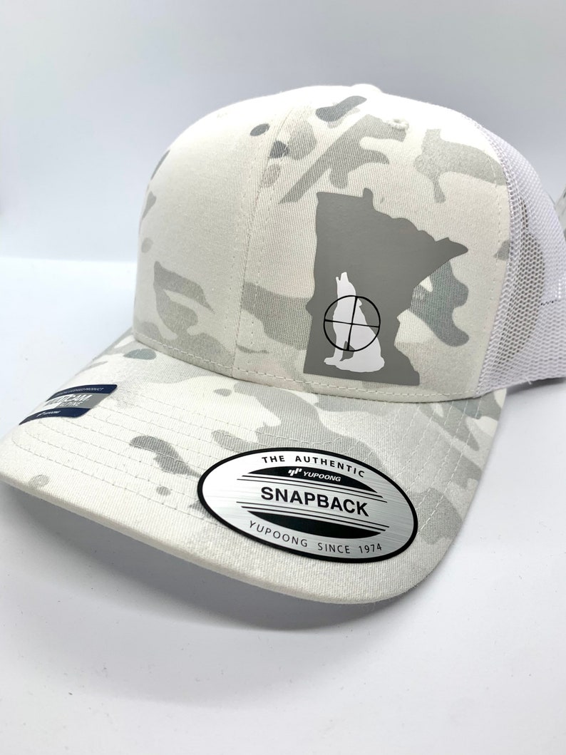 Pode incluir: Bon&eacute; de caminhoneiro branco camuflado com um contorno cinza de Minnesota e uma silhueta de lobo dentro de um alvo. O bon&eacute; tem uma malha branca na parte de tr&aacute;s e um adesivo ovalado prateado que diz "The Authentic Yupoong Snapback Yupoong Since 1974".