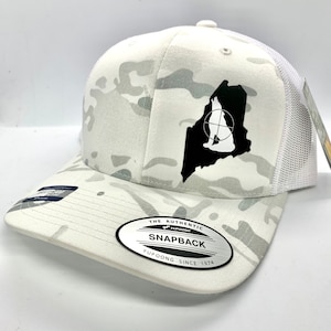 Pode incluir: Bon&eacute; de caminhoneiro branco camuflado com uma silhueta preta de um lobo uivando dentro do contorno do estado do Maine. O bon&eacute; tem um adesivo preto e branco na parte de tr&aacute;s que diz "The Authentic Yupoong Snapback Yupoong Since 1974."