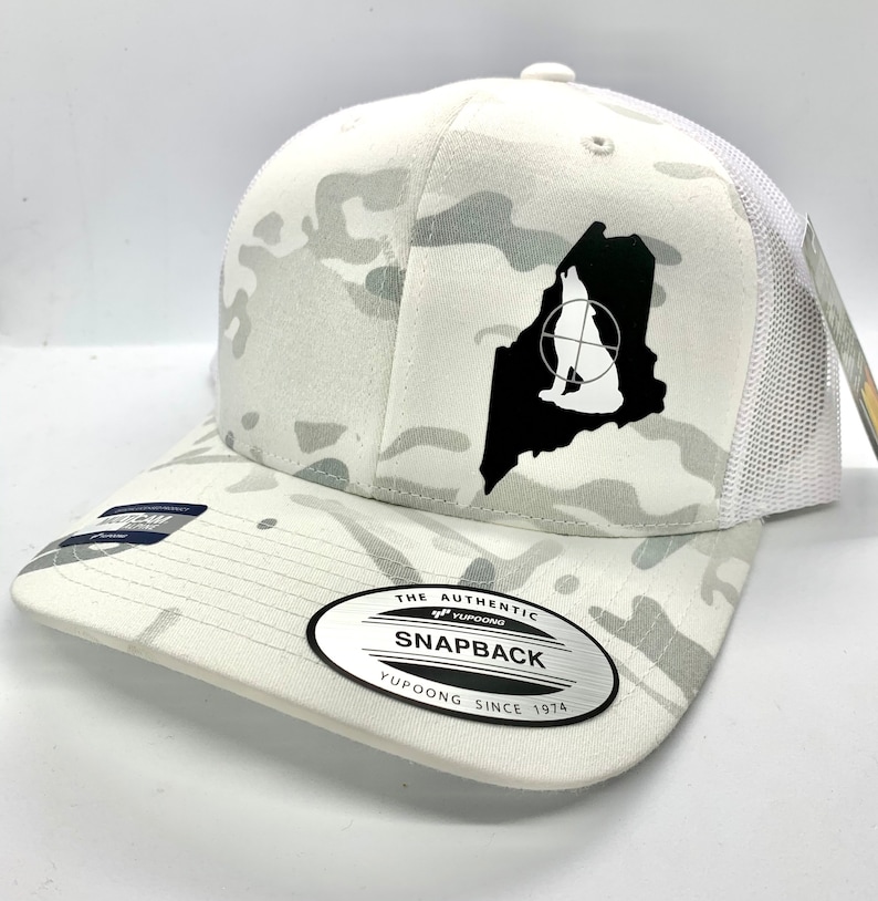 Pode incluir: Bon&eacute; de caminhoneiro branco camuflado com uma silhueta preta de um lobo uivando dentro do contorno do estado do Maine. O bon&eacute; tem um adesivo preto e branco na parte de tr&aacute;s que diz "The Authentic Yupoong Snapback Yupoong Since 1974."
