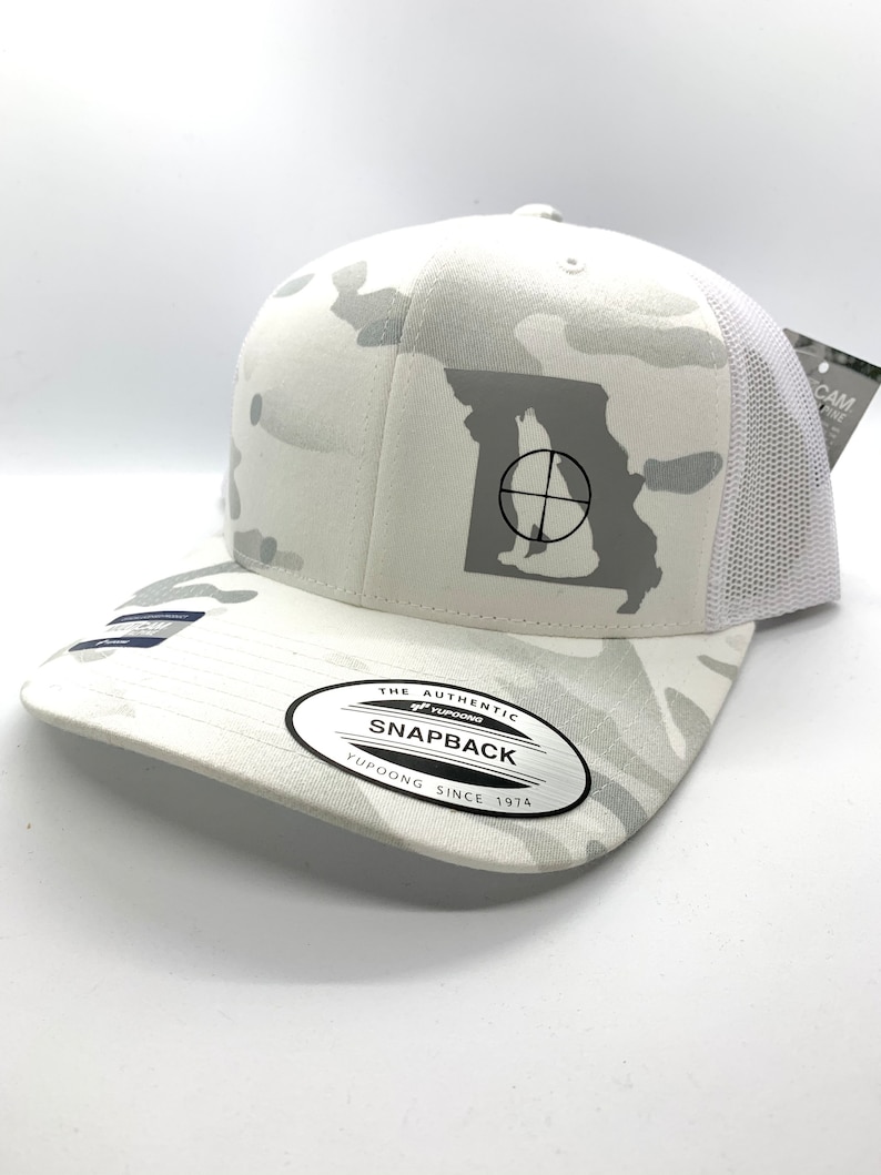 Pode incluir: Bon&eacute; de caminhoneiro branco camuflado com um contorno branco do estado do Missouri com um s&iacute;mbolo de alvo no interior. O bon&eacute; tem uma etiqueta preta "Yupoong The Authentic Snapback Yupoong Since 1974" na parte inferior da aba.