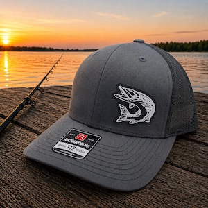 Puede incluir: Gorra de camionero gris Richardson 112 con un diseño de pez blanco. La gorra tiene una parte trasera de malla y una correa ajustable. La gorra está sobre una superficie de madera con una caña de pescar y un lago al fondo.