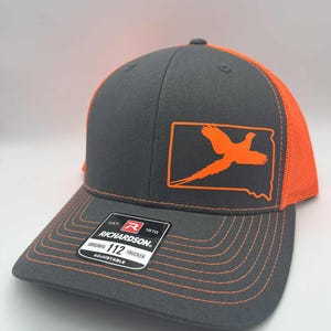 May include: A gray and orange Richardson 112 Trucker hat with an orange pheasant graphic inside a state outline. The hat has a curved brim and adjustable strap. The text "Original 112 Trucker Adjustable" is visible on a tag.
