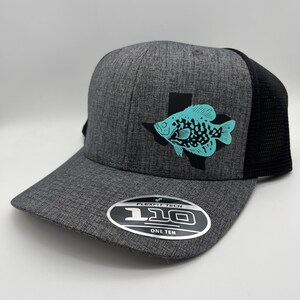 May include: A gray Flexfit baseball cap with a black mesh back. The cap features a teal fish graphic inside a black Texas outline. The Flexfit label is visible on the brim.