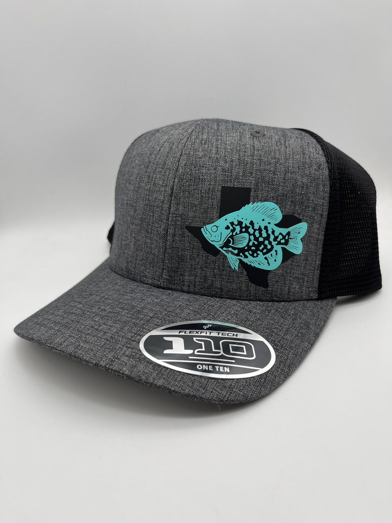 May include: A gray Flexfit baseball cap with a black mesh back. The cap features a teal fish graphic inside a black Texas outline. The Flexfit label is visible on the brim.