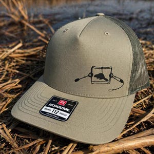 May include: An olive green trucker hat with a mesh back and a solid front panel. A black graphic of a beaver inside a square frame with a tethered trap is printed on the front. A black and white sticker on the brim reads Richardson 112 Trucker.
