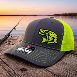 May include: A gray Richardson 112 trucker hat with a neon green fish graphic. The hat has a neon green mesh back and stitching. The hat is adjustable.