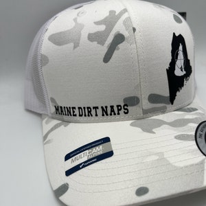 Pode incluir: Bon&eacute; de caminhoneiro branco camuflado com um contorno preto do estado do Maine e as palavras "Maine Dirt Naps" na frente. O bon&eacute; tem uma etiqueta "Multicam Alpine" preta e branca no lado.