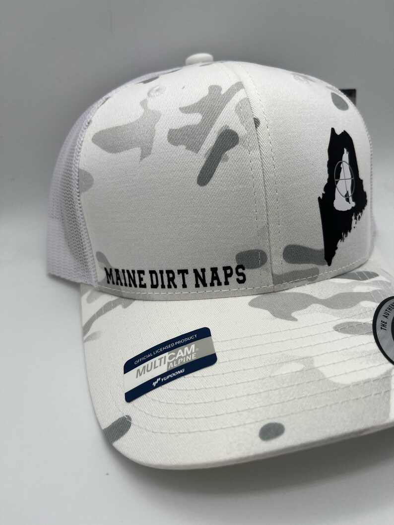 Pode incluir: Bon&eacute; de caminhoneiro branco camuflado com um contorno preto do estado do Maine e as palavras "Maine Dirt Naps" na frente. O bon&eacute; tem uma etiqueta "Multicam Alpine" preta e branca no lado.
