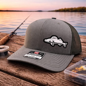 May include: A gray Richardson 112 Trucker hat with a white fish patch. The hat has a mesh back and an adjustable closure. The hat is sitting on a wooden dock with fishing gear in the background.