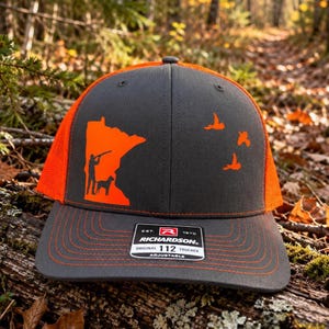 Puede incluir: Una gorra de camionero gris carbón y naranja brillante con la silueta del estado de Minnesota, un cazador, un perro y tres aves volando. El panel frontal muestra el texto Richardson Original 112 Adjustable.