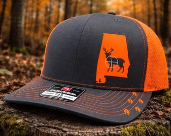 May include: A charcoal gray and orange trucker hat featuring the outline of Alabama in orange with a deer silhouette and crosshairs. The hat has a mesh back and a curved brim with orange stitching and deer tracks.