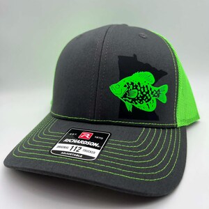 May include: A gray trucker hat with a neon green fish graphic and a black state outline. The hat has a neon green mesh back and stitching. The Richardson brand label is visible on the front.