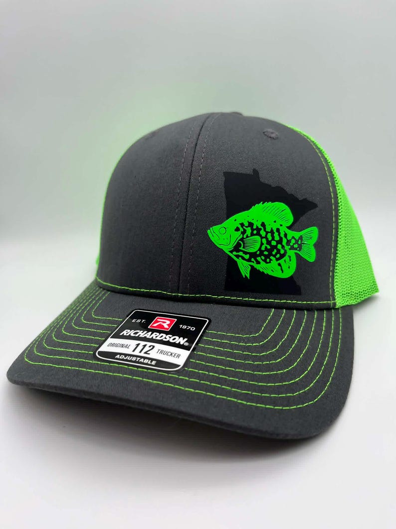 May include: A gray trucker hat with a neon green fish graphic and a black state outline. The hat has a neon green mesh back and stitching. The Richardson brand label is visible on the front.
