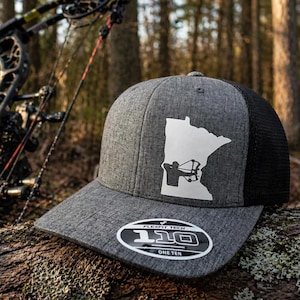 May include: A gray Flexfit baseball cap with a black mesh back. The cap features a white outline of a state with a bow hunter graphic. The brim has a Flexfit Tech 110 One Ten label.