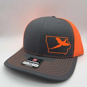 May include: Gray and orange trucker hat featuring an orange outline of the state of Iowa with a pheasant graphic. The hat has a curved brim, orange mesh back, and adjustable snap closure. The Richardson brand label is visible.
