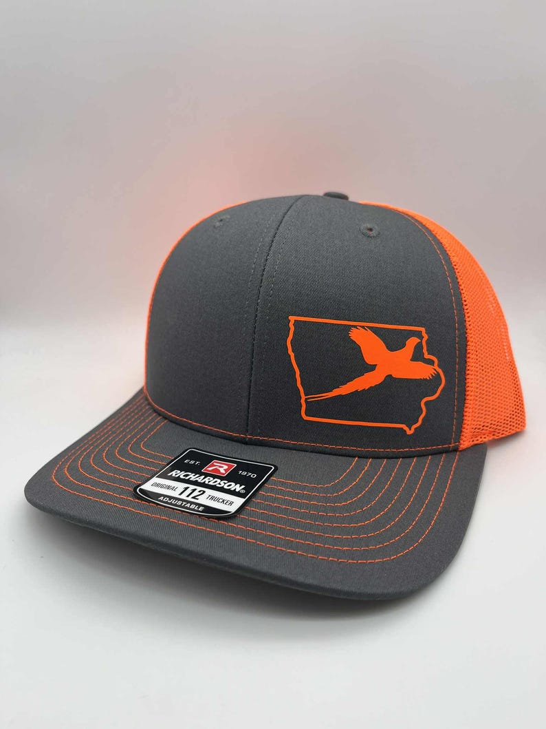 May include: Gray and orange trucker hat featuring an orange outline of the state of Iowa with a pheasant graphic. The hat has a curved brim, orange mesh back, and adjustable snap closure. The Richardson brand label is visible.