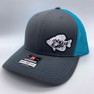 May include: A gray Richardson 112 trucker hat with a teal mesh back and brim stitching. A white and black fish graphic is on the front. The hat is adjustable.