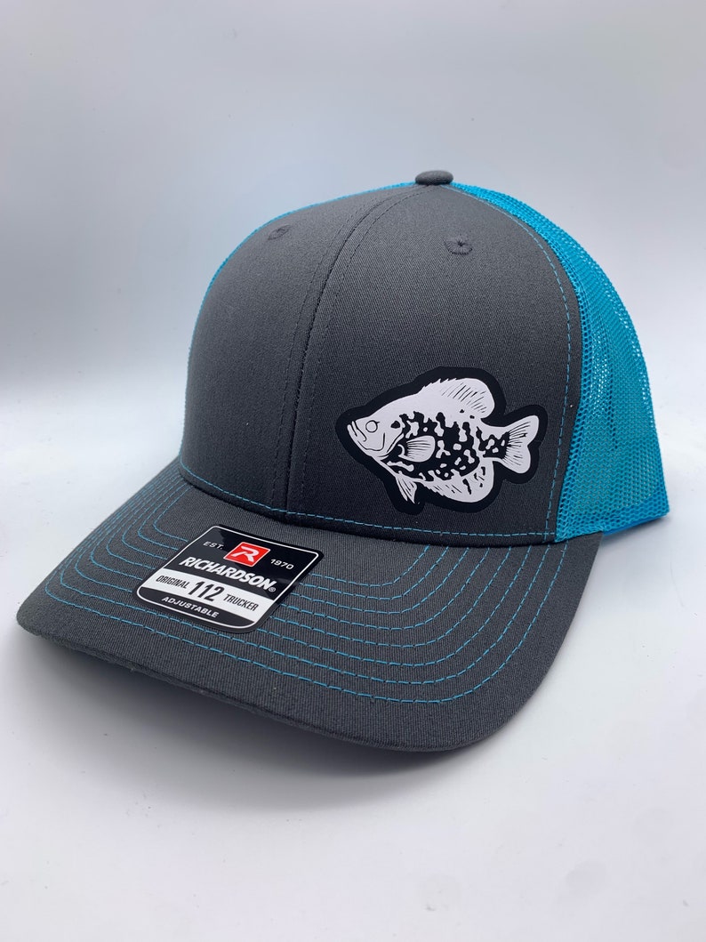 May include: A gray Richardson 112 trucker hat with a teal mesh back and brim stitching. A white and black fish graphic is on the front. The hat is adjustable.