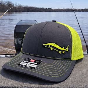 May include: Charcoal gray and neon yellow trucker hat with a fish emblem. The hat features a structured front panel, a curved brim, and a mesh back. The fish emblem is a bright yellow color. The hat is adjustable and has the Richardson brand label.