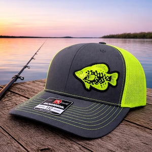 May include: A gray and neon green trucker hat with a fish patch. The hat features a dark gray front panel with a neon green fish graphic and a neon green mesh back. The brim and stitching are also neon green. The hat is adjustable.