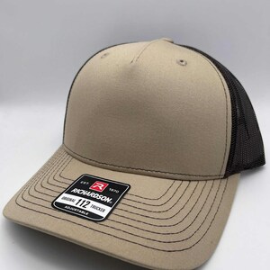 May include: A tan and black trucker hat with a curved brim. The hat features a mesh back and a structured front panel. A black and white tag on the brim reads "Richardson Original 112 Trucker Adjustable."
