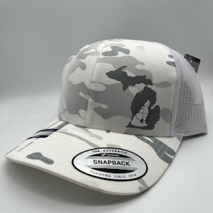 Pode incluir: Bon&eacute; de caminhoneiro de camuflagem branca com uma silhueta cinza de Michigan e um s&iacute;mbolo de alvo preto. O bon&eacute; tem uma parte de tr&aacute;s de malha branca e uma etiqueta preta "The Authentic Yupoong Snapback Yupoong Since 1974".