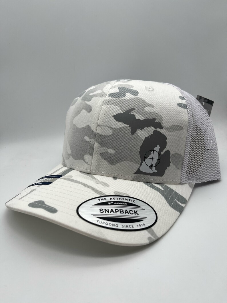 Pode incluir: Bon&eacute; de caminhoneiro de camuflagem branca com uma silhueta cinza de Michigan e um s&iacute;mbolo de alvo preto. O bon&eacute; tem uma parte de tr&aacute;s de malha branca e uma etiqueta preta "The Authentic Yupoong Snapback Yupoong Since 1974".