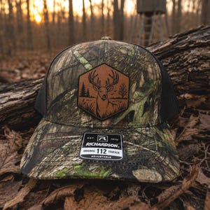 May include: A camouflage trucker hat with a black mesh back and a brown hexagonal leather patch featuring an engraved deer and forest scene. A black rectangular sticker on the brim reads EST. 1970 RICHARDSON ORIGINAL 112 TRUCKER ADJUSTABLE.