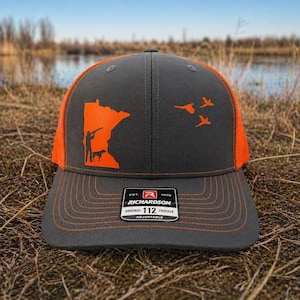 Może przedstawiać: Szara i pomarańczowa czapka typu trucker z grafiką myśliwego i psa na tle zarysu stanu. Czapka ma siatkowy tył i napis "RICHARDSON ORIGINAL 112 TRUCKER ADJUSTABLE".