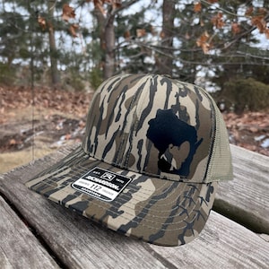 May include: Camouflage pattern trucker hat with a black silhouette of a state and a hunter. The hat has a mesh back and a curved brim. The tag reads "RICHARDSON 112 TRUCKER ADJUSTABLE".