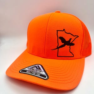 May include: Bright orange baseball cap with a black graphic of a pheasant inside a state outline. The cap has a mesh back and a curved bill. The label on the bill reads "Pacific Headwear Built For You."