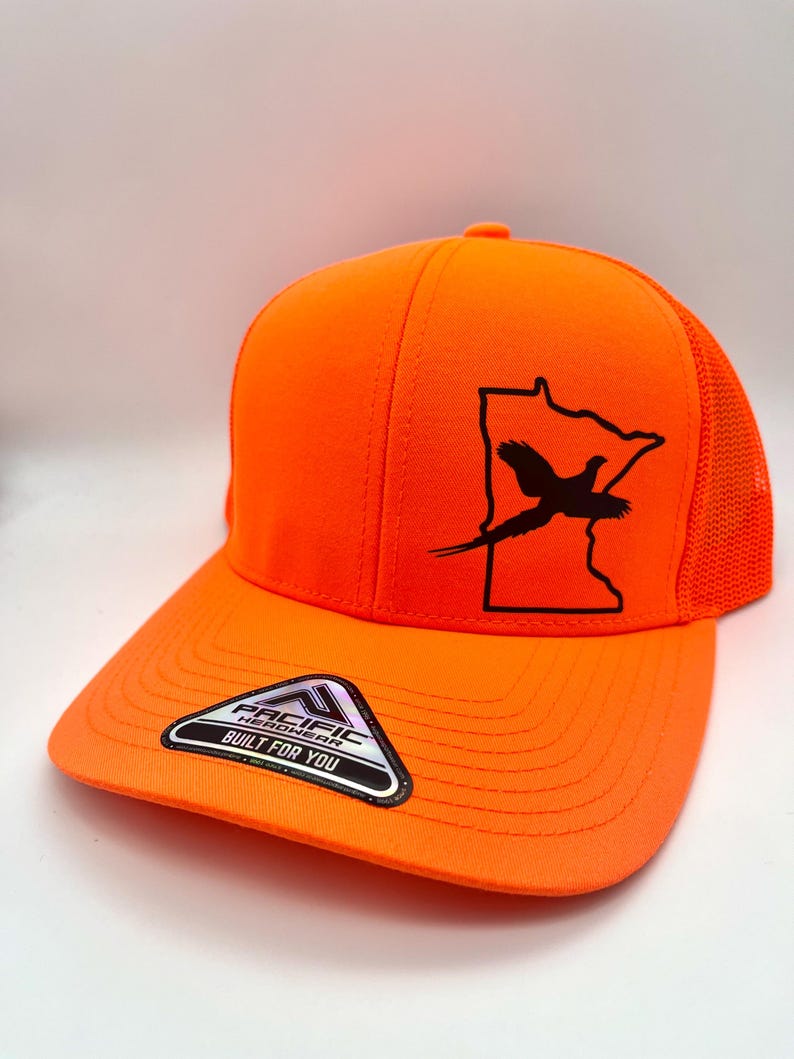 May include: Bright orange baseball cap with a black graphic of a pheasant inside a state outline. The cap has a mesh back and a curved bill. The label on the bill reads "Pacific Headwear Built For You."