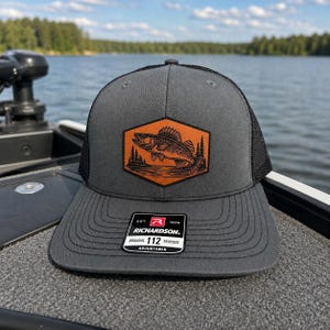 May include: Gray and black Richardson 112 trucker hat with a fish and tree patch. The patch is a brown hexagon with a fish and tree design. The hat has a curved brim and an adjustable strap.