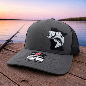 May include: A charcoal gray trucker hat with a black mesh back. The hat features a white and black graphic of a fish jumping out of the water, set against a black outline of a state. The hat has a curved brim and an adjustable strap.