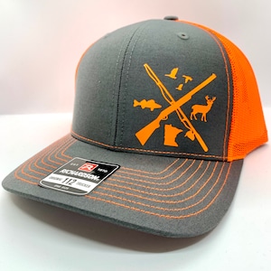 Puede incluir: Una gorra de camionero gris y naranja con un diseño de temática de caza. El diseño presenta un rifle y una escopeta cruzados, un ciervo, un pez y un pájaro. La gorra tiene una etiqueta negra con el texto "EST. 1970 RICHARDSON 112 TRUCKER ONE SIZE"