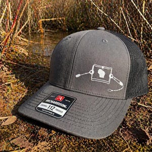 Puede incluir: Gorra de camionero gris con parte trasera de malla negra. La gorra presenta un gráfico blanco de un mapa y equipo de pesca. La visera tiene una etiqueta en blanco y negro que dice "RICHARDSON" y "112 TRUCKER".