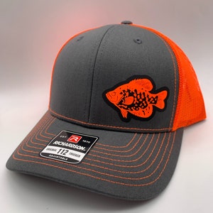 May include: A gray trucker hat with an orange fish graphic on the front. The hat has an orange mesh back and bill with orange stitching. The Richardson brand label is visible on the front.