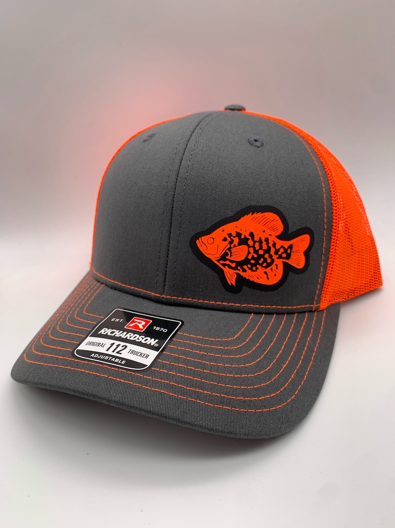 May include: A gray trucker hat with an orange fish graphic on the front. The hat has an orange mesh back and bill with orange stitching. The Richardson brand label is visible on the front.