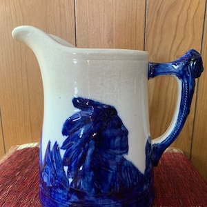 May include: A white ceramic pitcher with a blue and white design of a Native American man in profile. The pitcher has a handle shaped like a Native American head.
